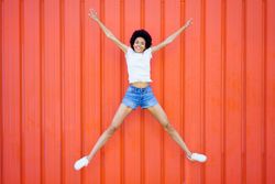 Woman in shorts and t-shirt jumping for joy against red wall