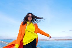Trendy woman in sunglasses, orange coat, yellow hoodie and jeans in front of the sea