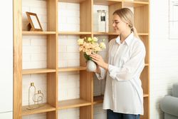 Young woman with flowers near wooden shelves in modern flat