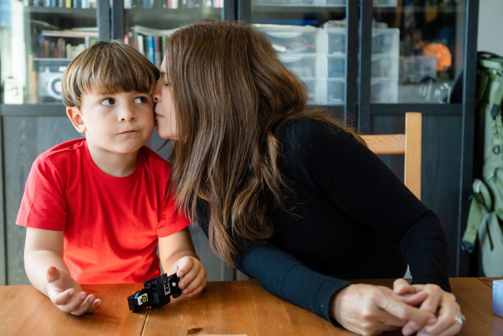 Loving mom kissing cute son on cheek while sitting at table