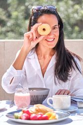 Positive woman looking through hole in donut while having breakf