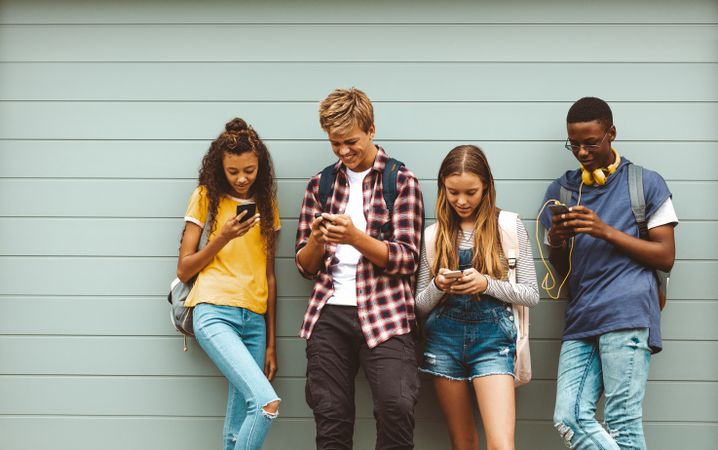 Group of teenage boys and girls standing outdoors against a wall looking at their phones