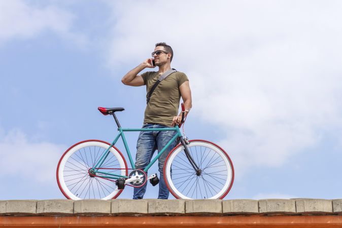 Male speaking on phone and standing with bike on roof with beautiful blue sky and clouds, copy space