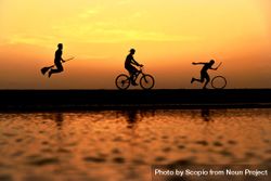 Silhouette of person riding witches broom and person riding a bicycle and another person moving a wheel near body of water during sunset 4mGAv0