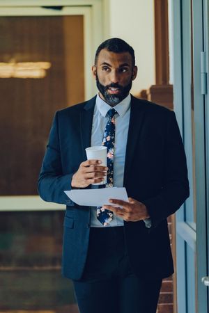 Serious businessman in suit and tie walking in office with to go coffee and paper