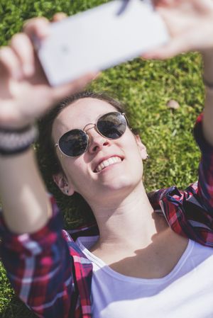 Above view of a young smiling hipster woman lying on grass in a sunny day at a park while taking a selfie with a mobile phone