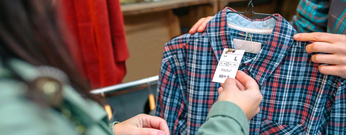 Banner of unrecognizable female customer looking label price of plaid shirt on second hand store