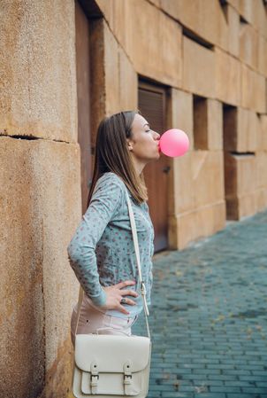 Side view of woman standing outside in front of stone wall blowing bubble gum with hands on hip