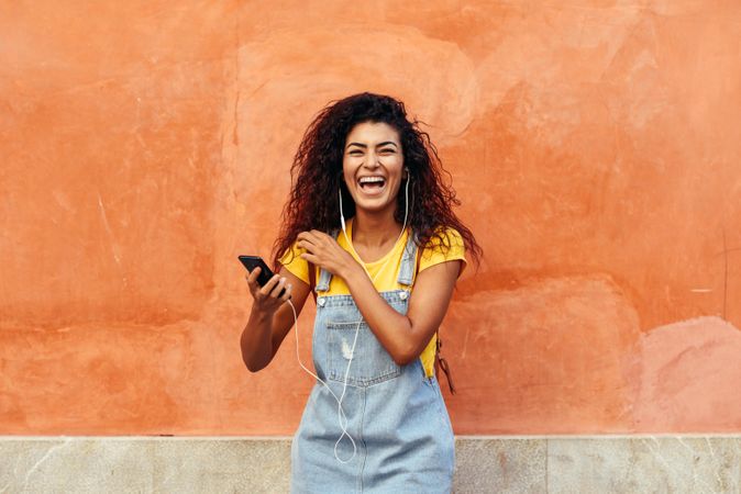 Laughing Arab woman with curly hairstyle using phone and headphones in front of on orange wall