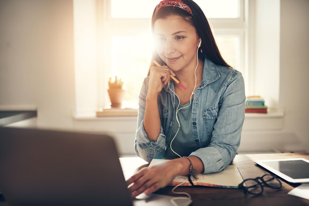 Content woman reading on laptop in home office with earbuds and pencil