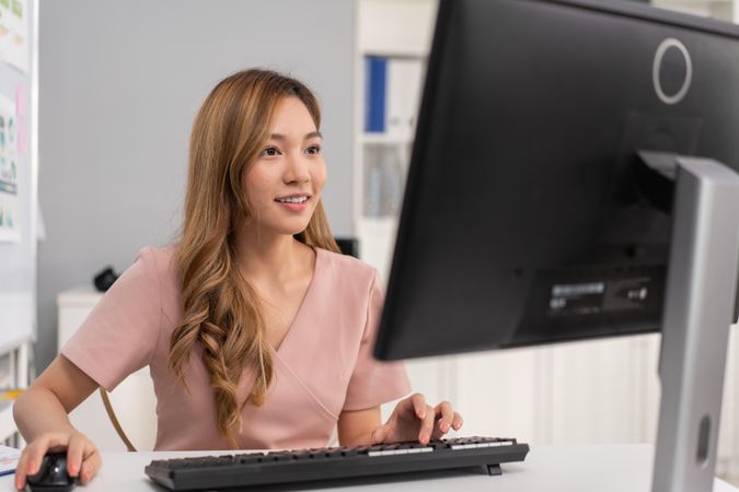 Confident Asian woman sitting in office and typing on keyboard while looking at monitor