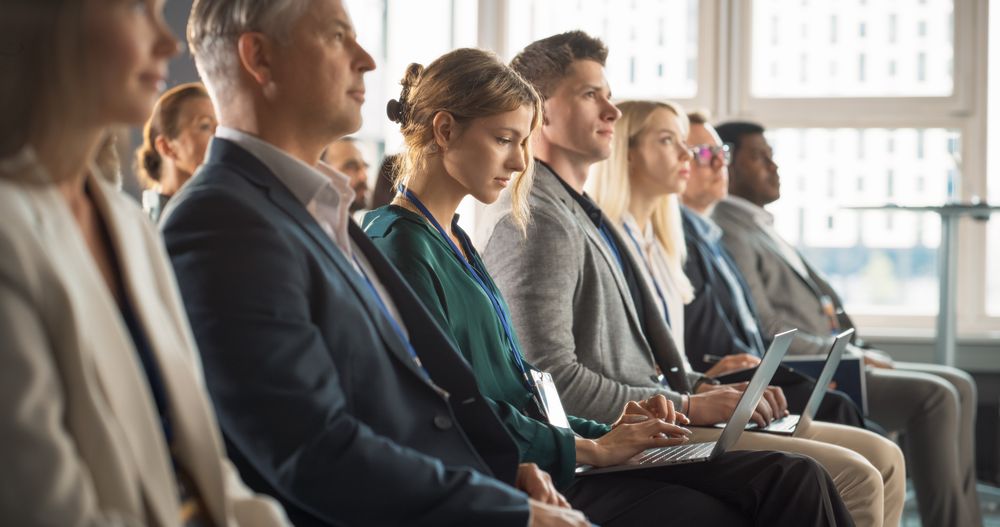 Young Woman Sitting in Crowded Audience at a Business Conference. Female Delegate Smiling and Using Laptop Computer. Manager Watching Motivational Presentation About Investing In Innovative Startups.