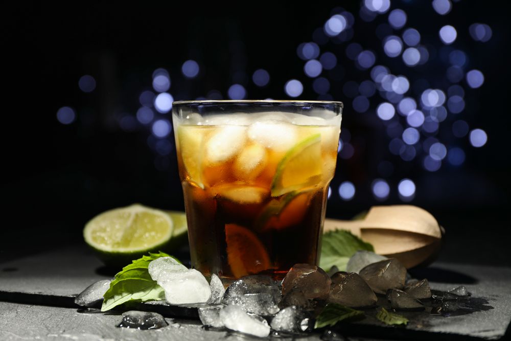 Glass with cola and lime slices, juicer, ice, mint against blurred lights background