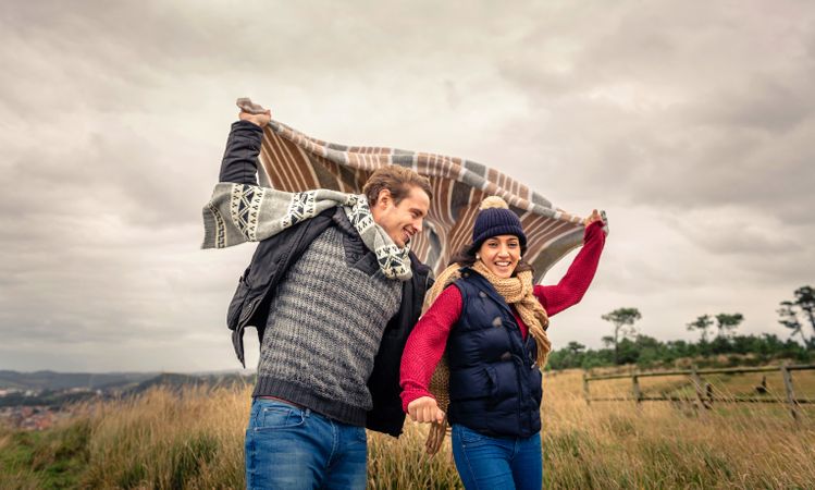 Portrait of young couple playing outdoors with blanket on a windy day over dark cloudy sky