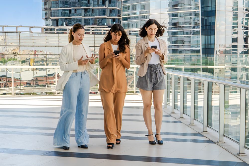 Three businesswomen are walking while using smartphones outdoors. All women are dressed in business attire..