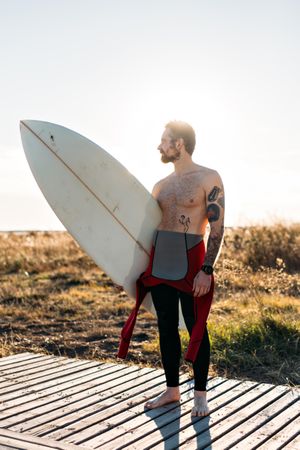Tattooed man in wetsuit standing on boardwalk leading to the beach with surfboard at sunset