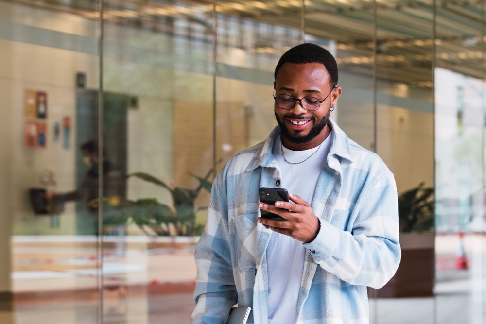 Cheerful Black man browsing smartphone near building