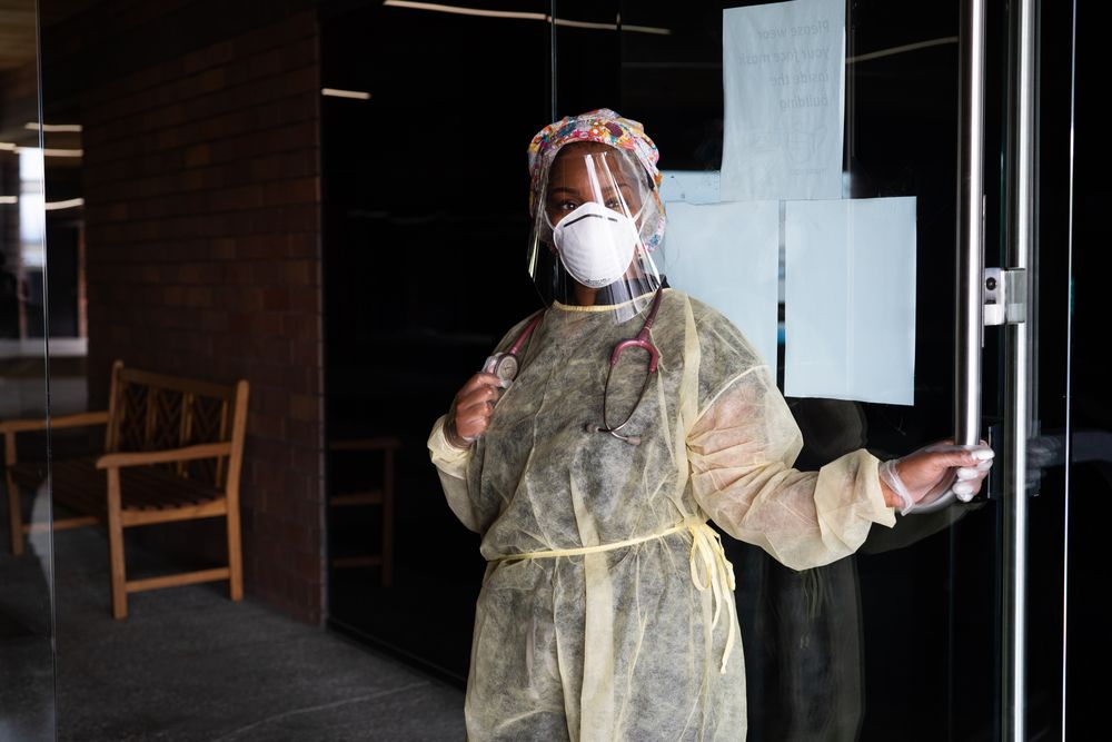 Nurse with PPE and stethoscope holding door of medical building open