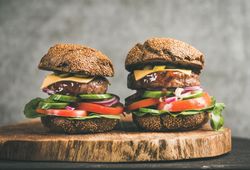 Two cheeseburgers, stacked with fresh vegetables, on wooden board