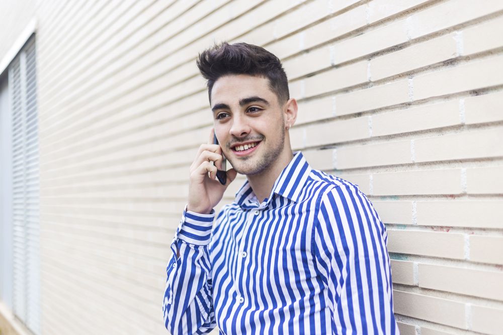 Man leaning on house wall talking on his smartphone and looking into the distance