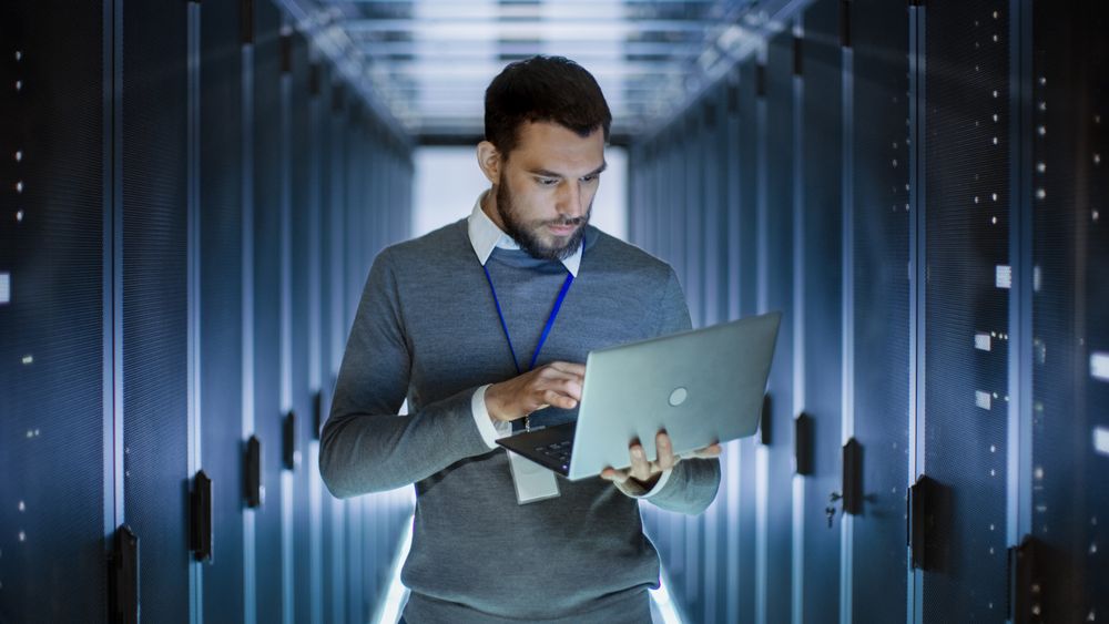 Male Server Engineer Works on a Laptop in Large Data Center.
