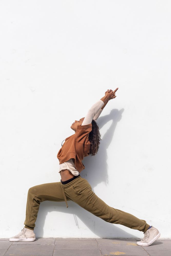 Stylish young female runner in athletic wear stretching before running against wall