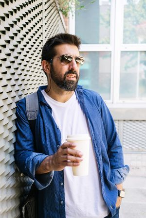 Latino man in denim leaning on fence outside with cup of coffee