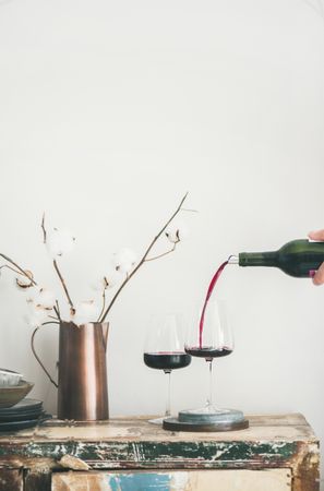Wine glasses being poured from bottle with dried cotton in vase, vertical composition, copy space