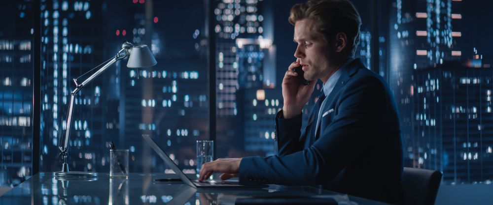 Successful Businessman Working on Laptop Computer And Picking Up Phone Call in Big City Office in the Evening.