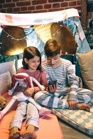 Smiling children looking mobile phone while chatting on social media application sitting on tent