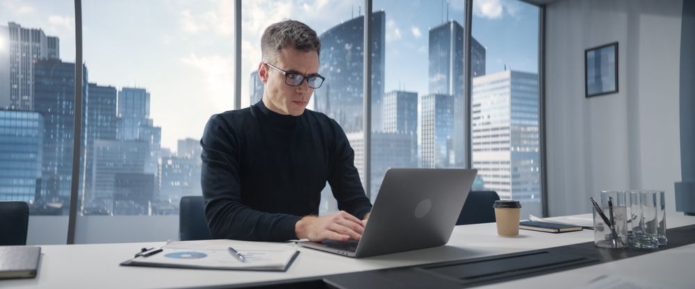Portrait of Successful Businessman Working on Laptop Computer in His Big City Corner Office.