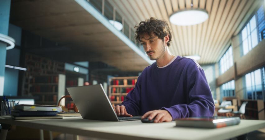 Student Studying in a Modern Library. Young Man Thinking and Problem Solving School Exercises. Male Using Laptop Computer to Work on a University Research Project Online