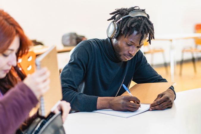 Black male student with headphones intently writing notes at a classroom desk, absorbed in study.
