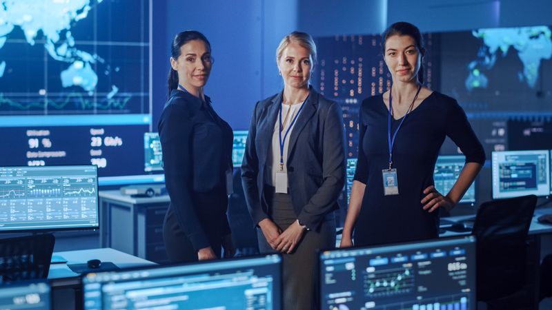 Three Professional Women Stand Confidently in a High-tech Control Room. Surrounded by Data Screens and World Maps, They Oversee Global Operations in a Blue-lit Environment.