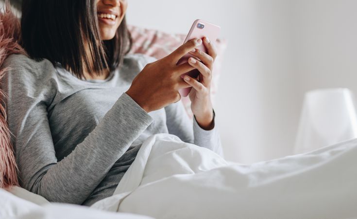 Cropped shot of smiling young woman sitting on bed checking her cell phone