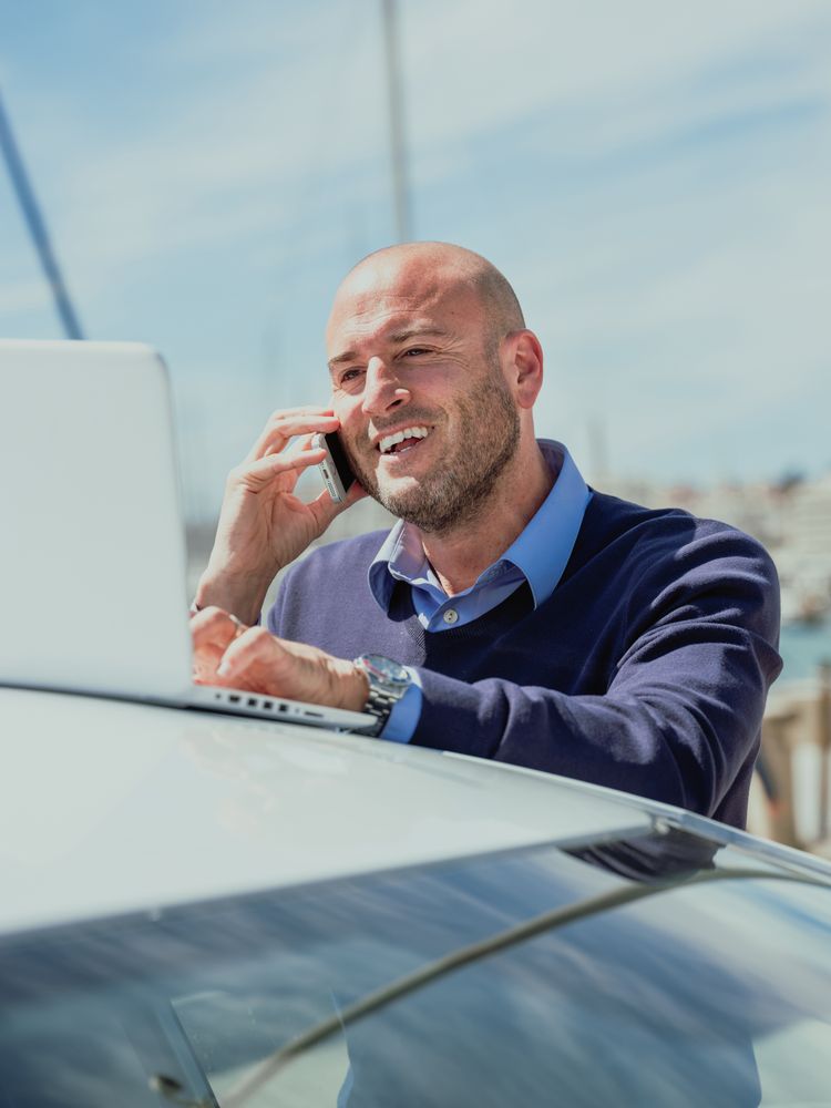 Vertical photo of a businessman talking on the phone while using a laptop supported by a car