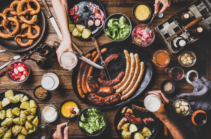 German sausages and pretzels displayed on wooden table with hands holding beer and serving food