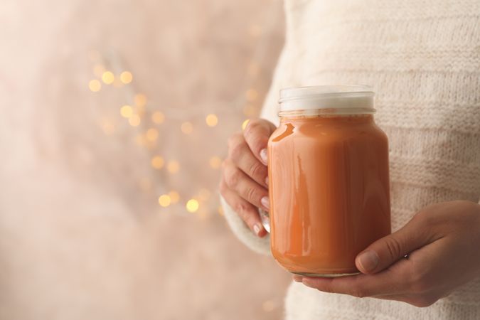 Woman hold glass jar of pumpkin latte against brown background with blurred lights