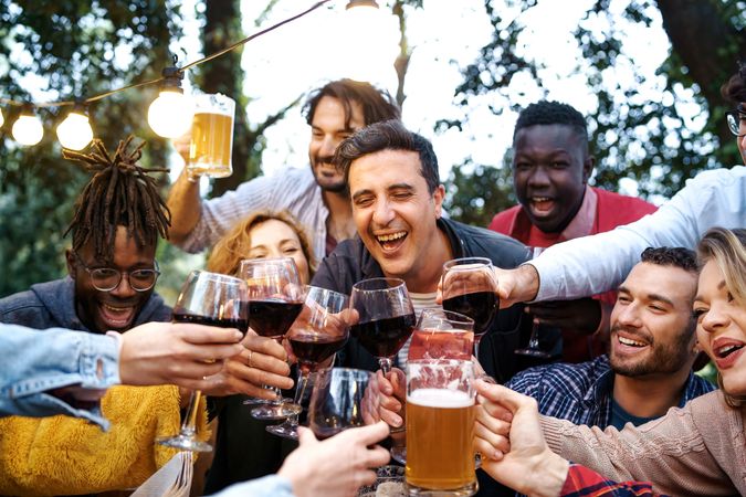 A diverse group of friends aged 20-40, toasting with beer, wine, and sangria glasses outdoors at a countryside house during dusk