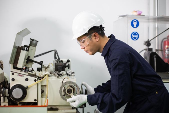 Asian man in hard hat and coveralls operating machine in factory