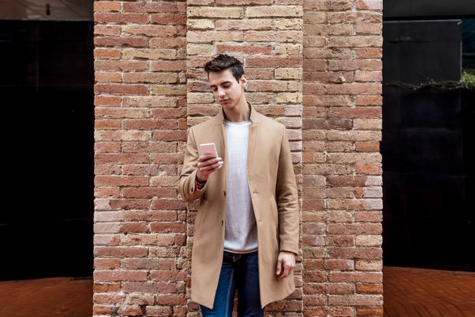 Front view of man in camel coat texting on phone while leaning on brick wall