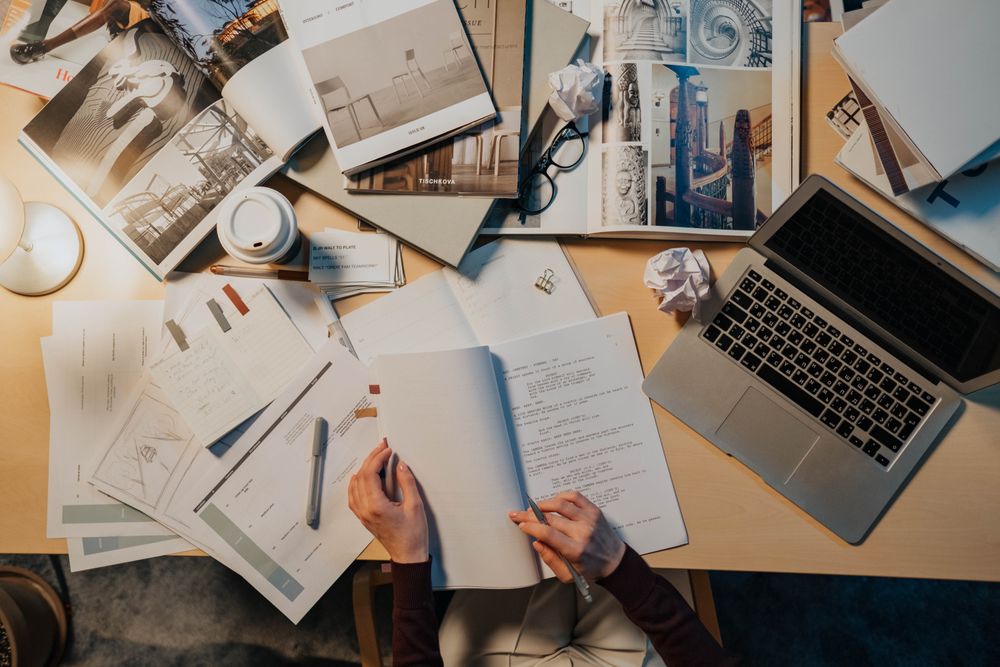 Top view of a person writing on a paper beside books and laptop on wooden desk