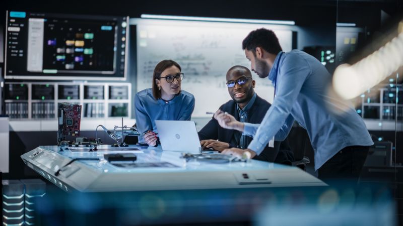 Diverse Team of Engineers Collaborating on Manufacturing Project in a Research Facility. Colleagues Analysing Data Using Laptop Computer and Brainstorming for a Technologically Advanced Invention