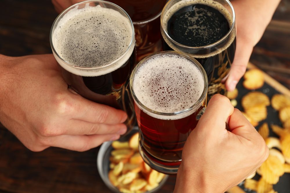 Four men cheers with beer on wooden background with snacks