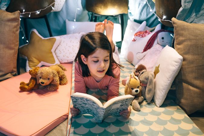 Portrait of serene little girl looking book while resting surrounded by cushions in homemade teepee