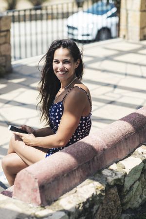 Above view of a smiling woman in blue dress sitting on a bench holding a mobile phone