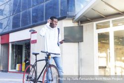 Young Black man walking in the street with bike and adjusting his headphones 0WOVJP