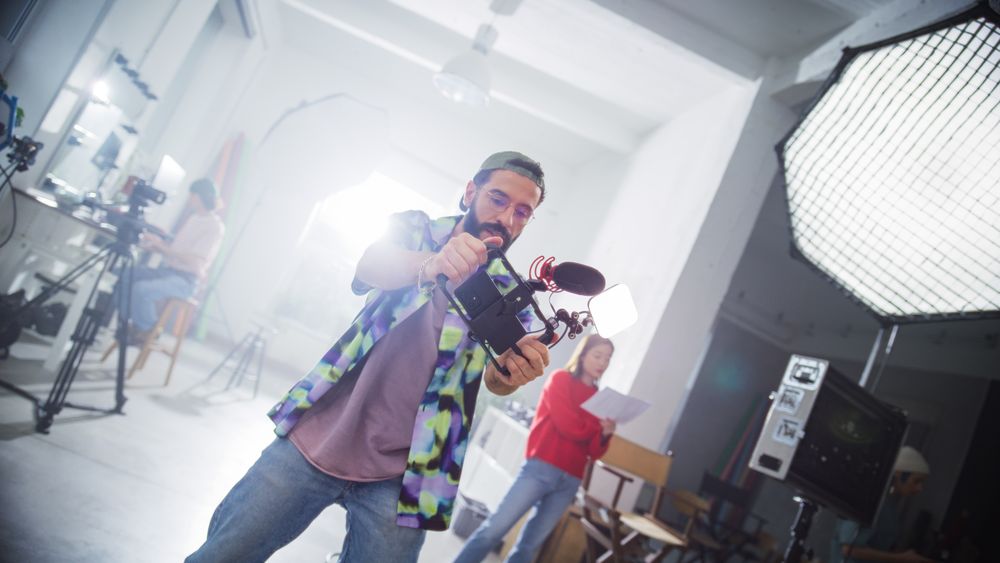 Focused Young Male Cinematographer With Beard, Wearing A Cap And Plaid Shirt, Actively Filming In A Professional Studio Setting, Surrounded By Modern Filming Equipment And Crew Members.
