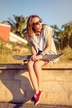 Young girl with skateboard sitting over the wall on summer