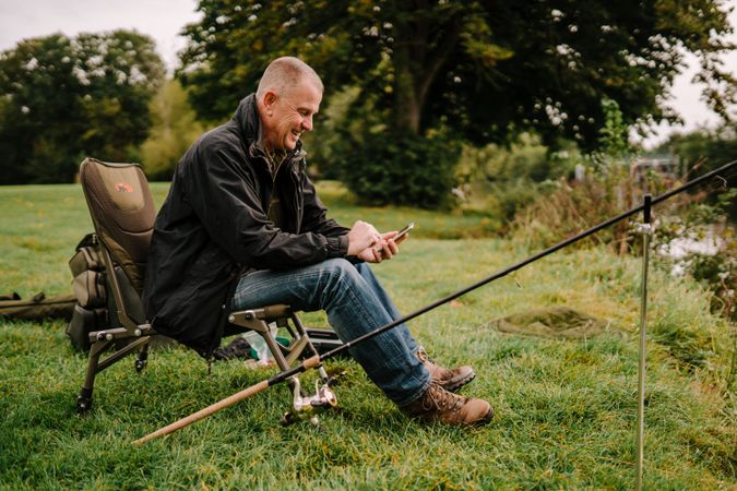 Happy sitting with fishing rod by river and checking phone